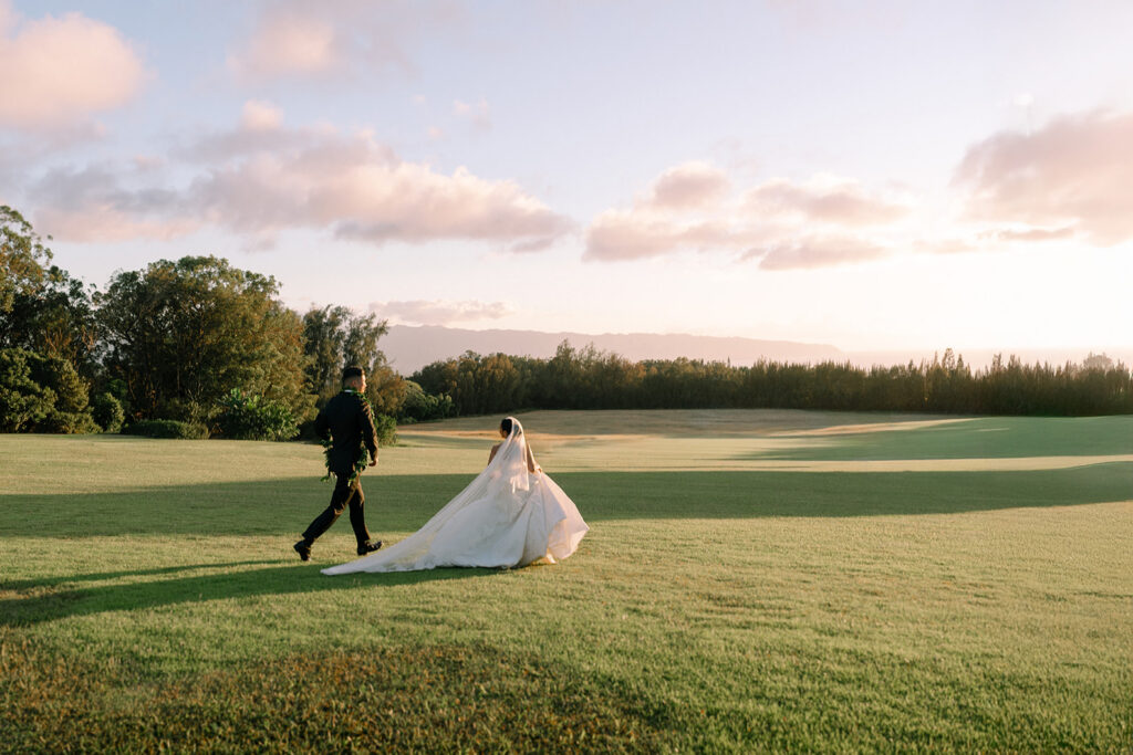 A bride and groom walk across a grassy field under a partly cloudy sky during sunset.