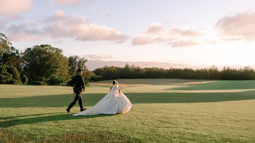 A bride and groom walk across a grassy field under a partly cloudy sky during sunset.