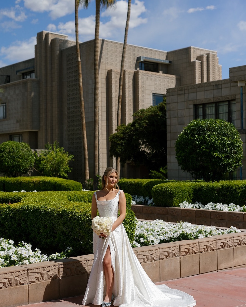 A bride in a white dress holding a bouquet stands outdoors in front of an art deco building, surrounded by landscaped greenery and flowers.