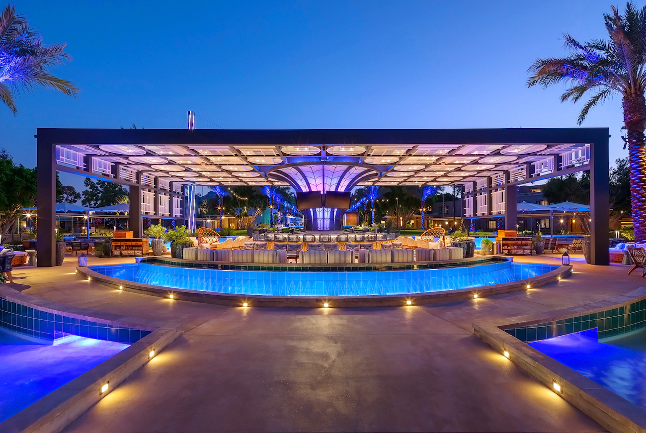 Outdoor bar and lounge area with illuminated pool, modern canopy roof, seating, and palm trees, photographed at dusk with blue accent lighting.