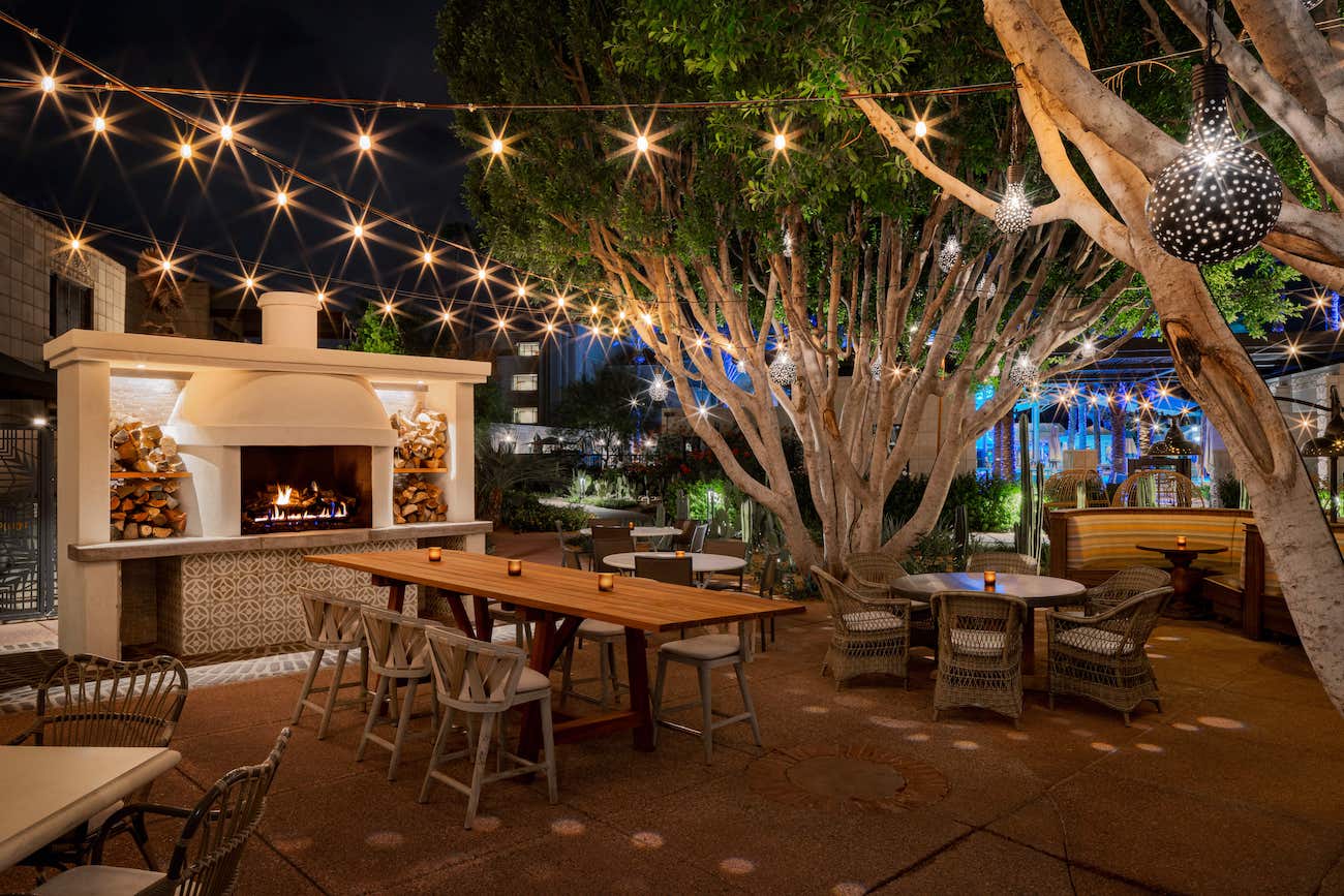 Outdoor patio at night with string lights, wooden tables, wicker chairs, and a lit fireplace surrounded by trees and modern decor.