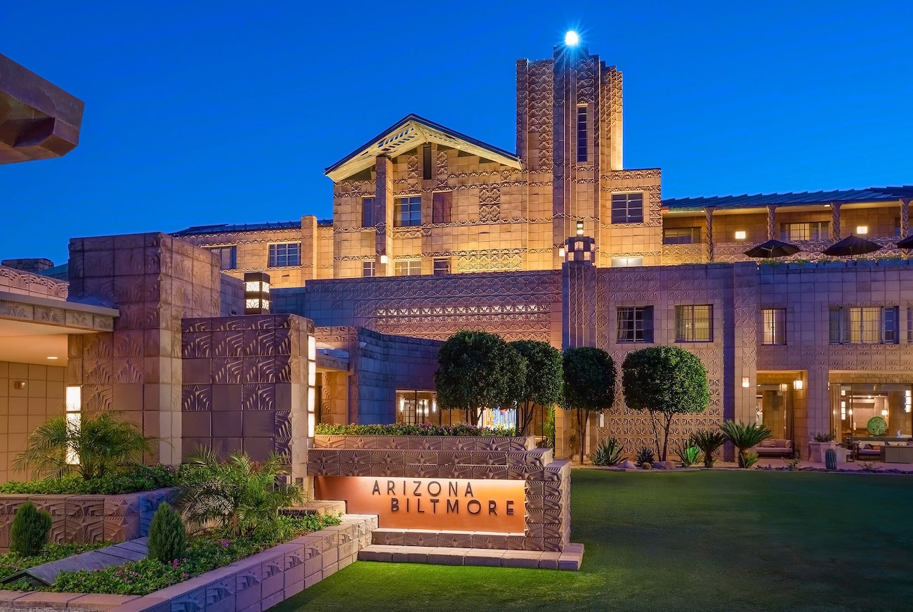 The Arizona Biltmore hotel exterior at dusk, with warm lights illuminating the building and a sign reading “Arizona Biltmore” in front.