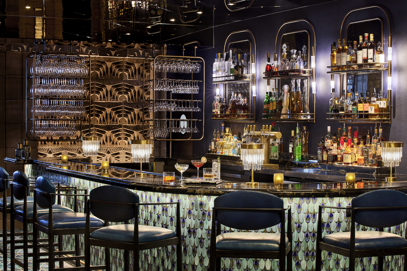 Elegant bar with a mirrored back wall, shelves of liquor bottles, glassware, bar stools, and glowing accent lighting.