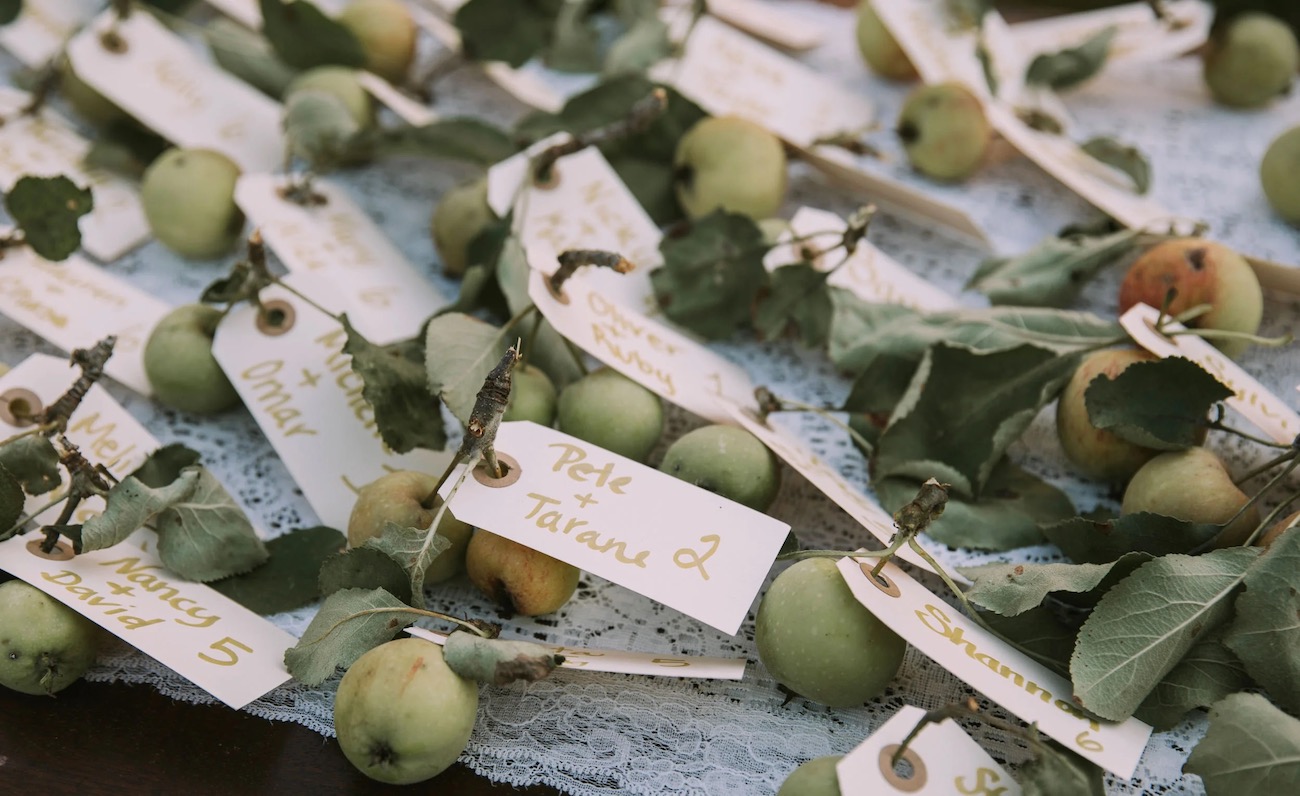 Small green apples with leaves are arranged on lace fabric, each bunch labeled with a handwritten tag for identification.