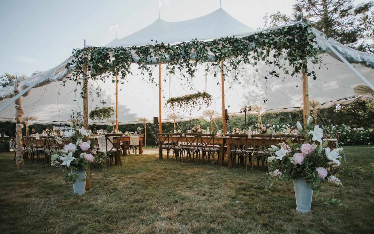 Outdoor wedding reception setup under a white tent with wooden tables, chairs, floral arrangements, and greenery decorations.