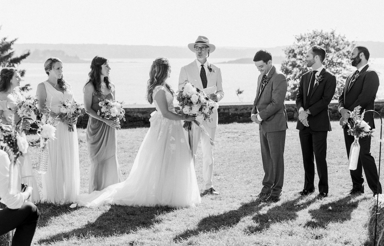 A bride and groom stand with their wedding party outdoors during a ceremony, with a person officiating. The scene is in black and white.