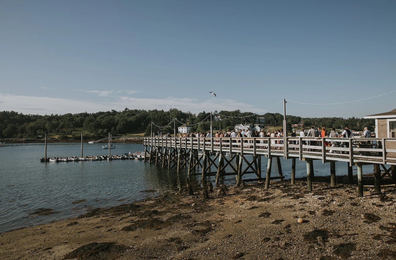 A wooden pier extends over calm water with people walking on it, surrounded by trees and buildings in the background under a clear sky.