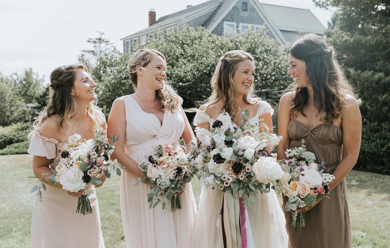Four women in light dresses stand outdoors holding bouquets, smiling at each other, with a house and greenery in the background.