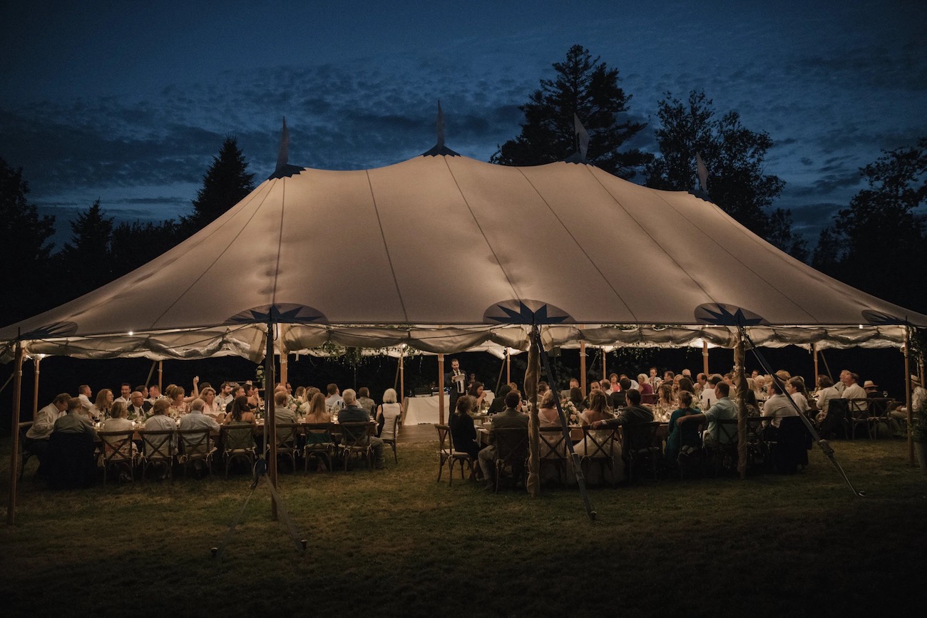 A large group of people sit at tables under a lit canopy tent outdoors at night, surrounded by trees.