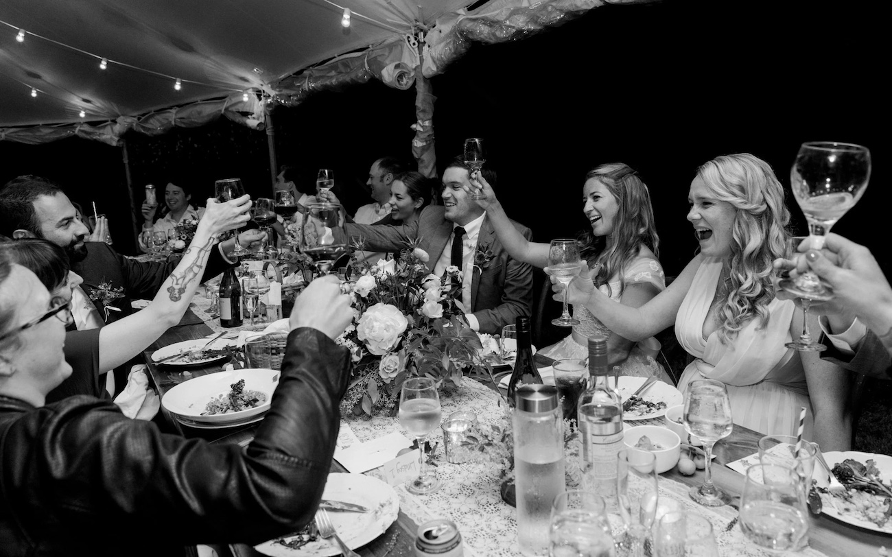 A group of people sit around a long table under string lights, raising their glasses in a toast at a festive gathering.