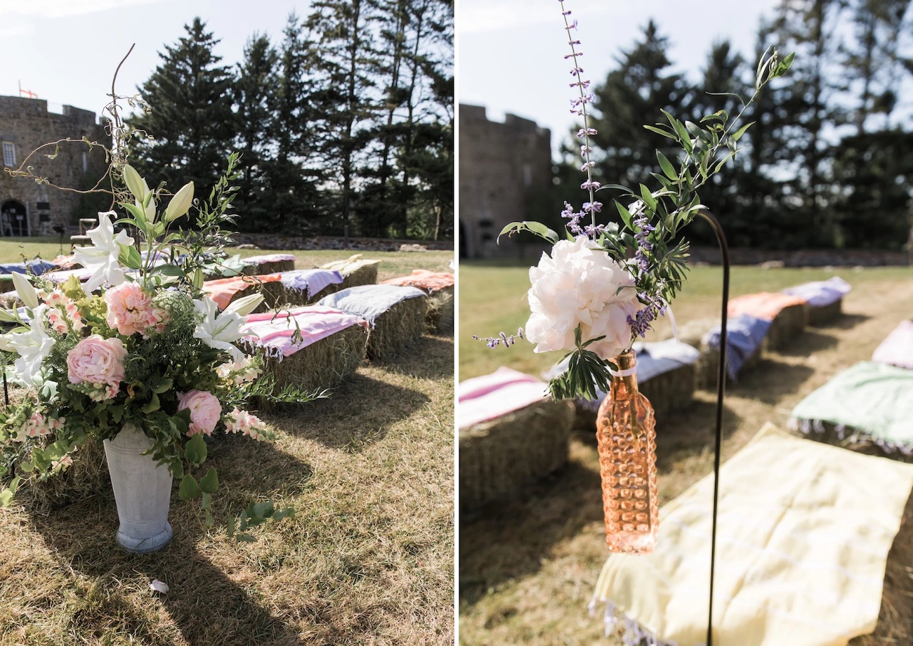 Outdoor wedding setup with hay bale seating, pastel blankets, and floral arrangements; trees and a stone building are visible in the background.