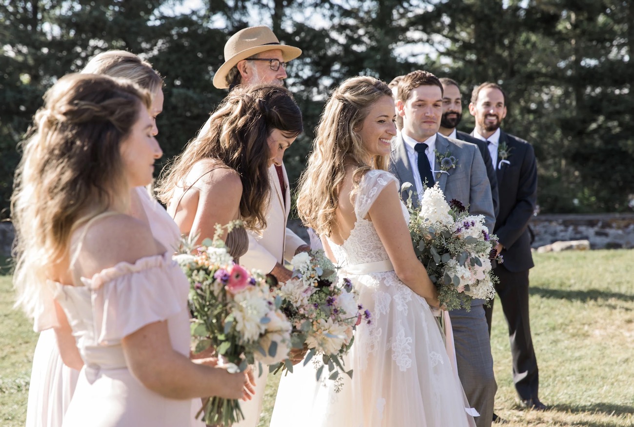 Bride and bridesmaids in light dresses and holding bouquets stand beside groom and groomsmen outdoors during a wedding ceremony.