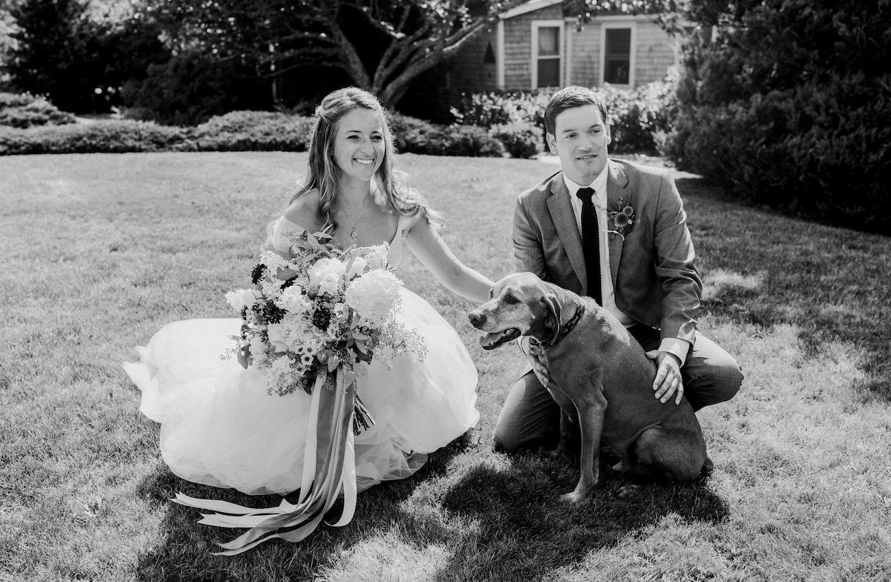 A bride and groom pose outdoors on grass with a large bouquet and a dog, with a house and trees in the background.