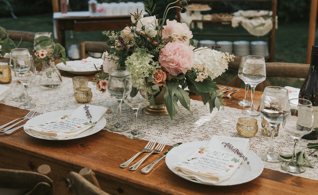 A rustic wooden table is set with floral centerpieces, plates, silverware, wine glasses, and lace runners for an outdoor event.