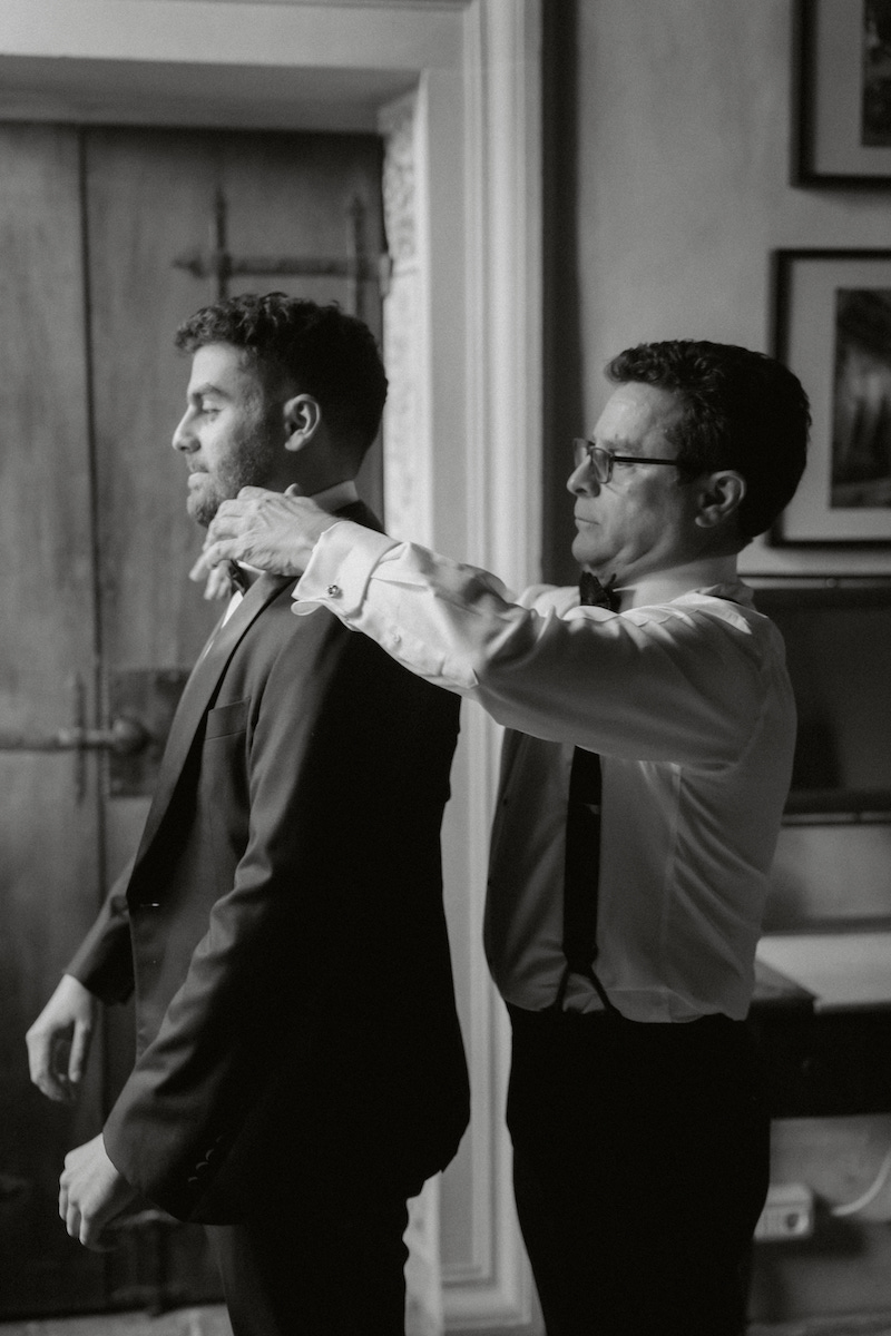 A man in suspenders adjusts the bow tie of another man wearing a suit in a room with framed pictures and a wooden door.