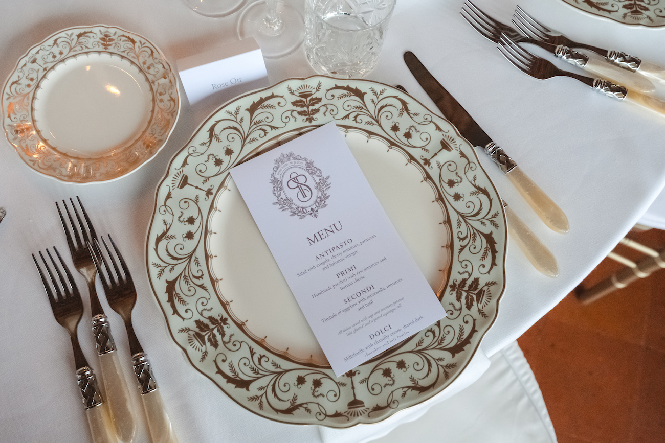 Elegant place setting with ornate plate, menu card, cutlery, glass, and side plate arranged on a white tablecloth at a formal event.