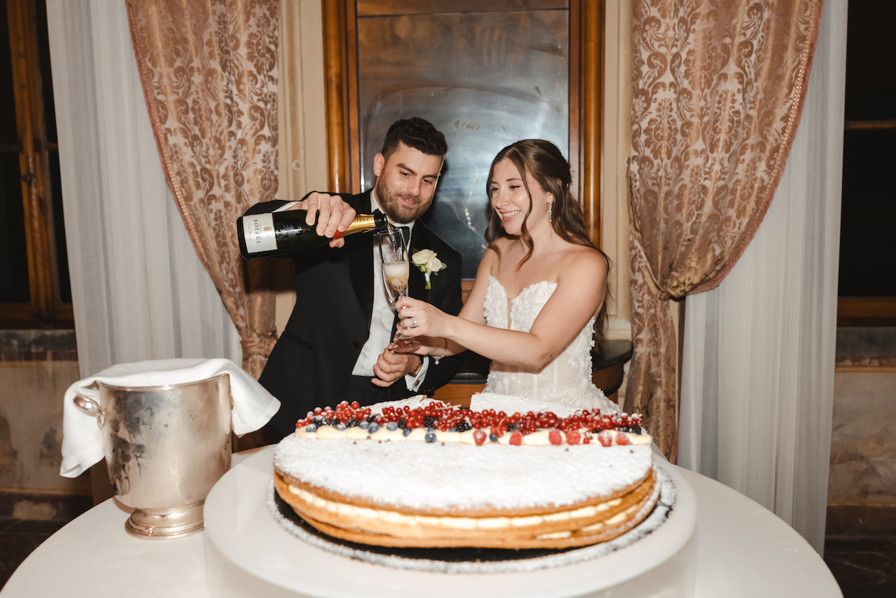 A bride and groom stand behind a large decorated cake as the groom pours champagne into the bride’s glass. They are indoors, with draped curtains and a window in the background.