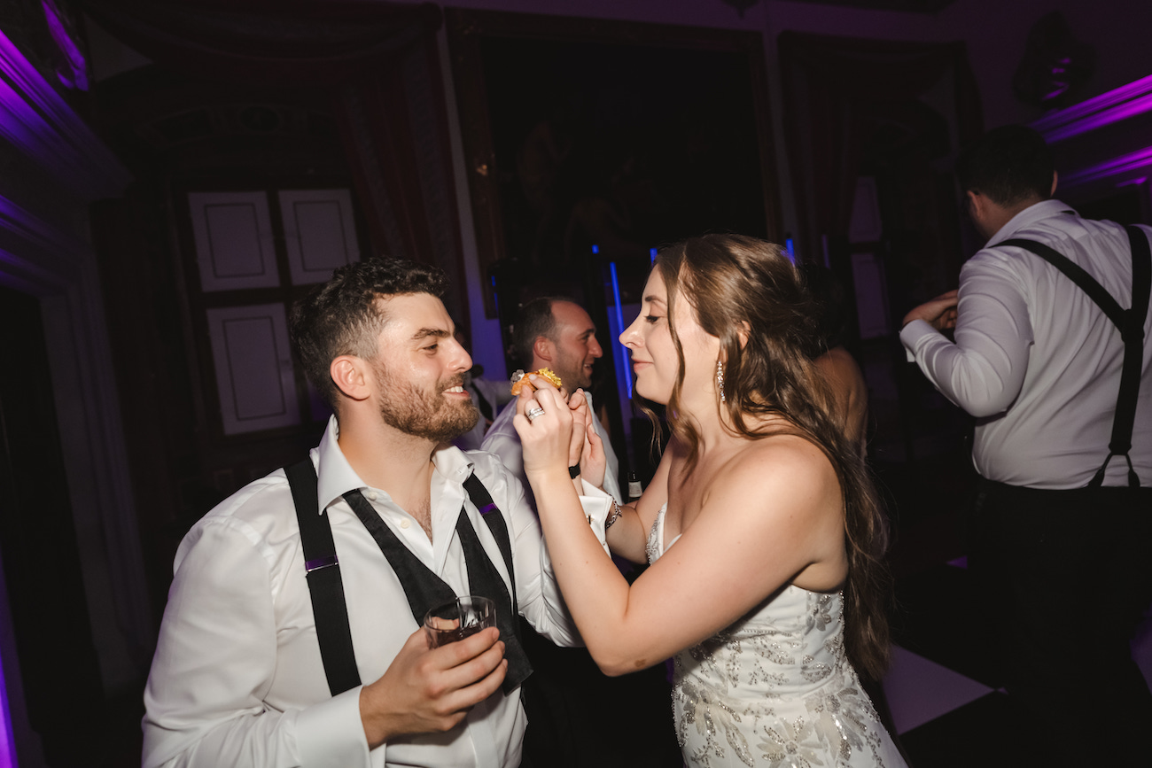 A man and woman in formal attire laugh and feed each other cake at a party, with other guests and dim lighting in the background.