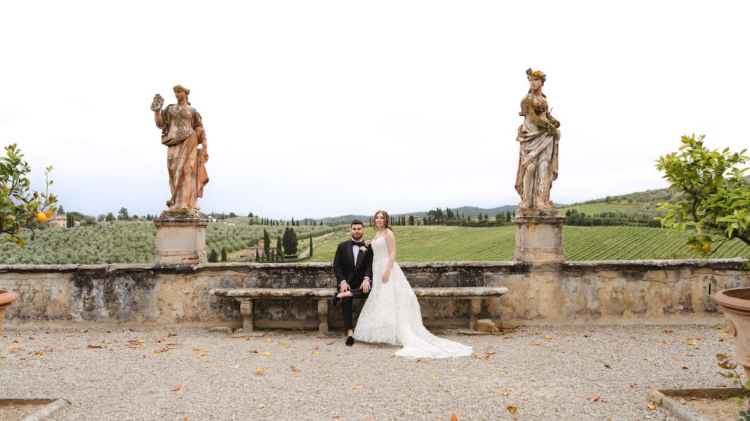 A bride and groom sit on a stone bench between two statues, with a scenic green landscape and cloudy sky in the background.