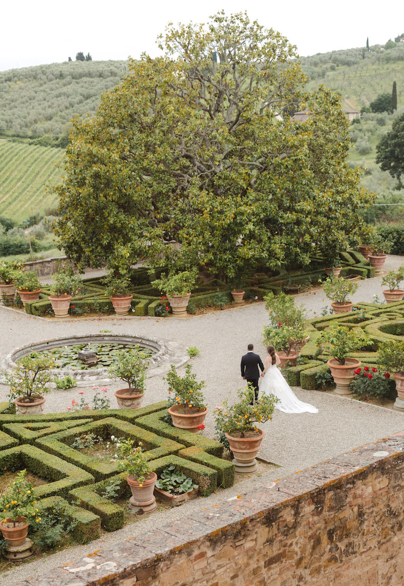 A bride and groom walk through a formal garden with geometric hedges, potted plants, and a large tree, surrounded by rolling green hills.