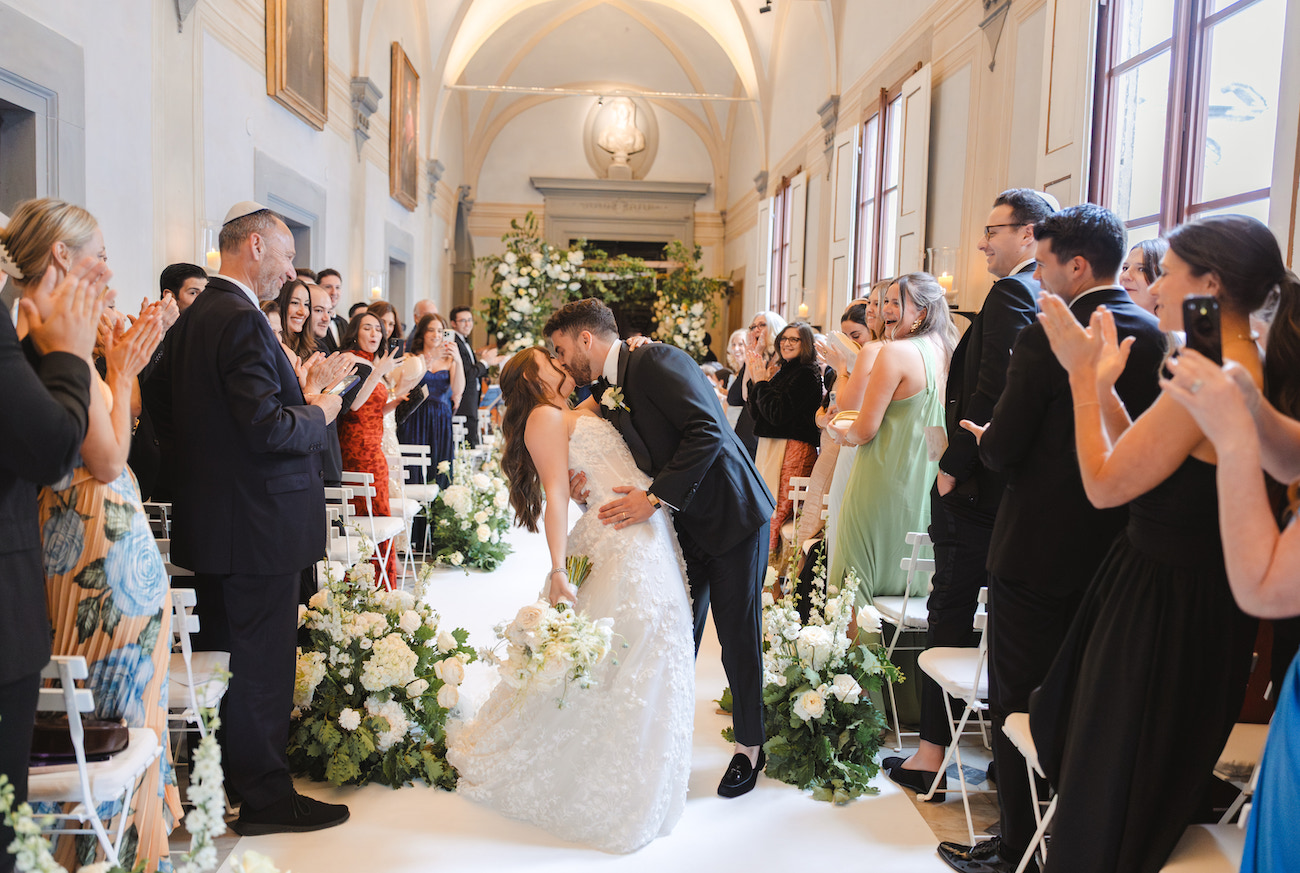 A bride and groom kiss at the end of an indoor wedding aisle, surrounded by guests who are standing, clapping, and smiling. The space is decorated with flowers and natural light.