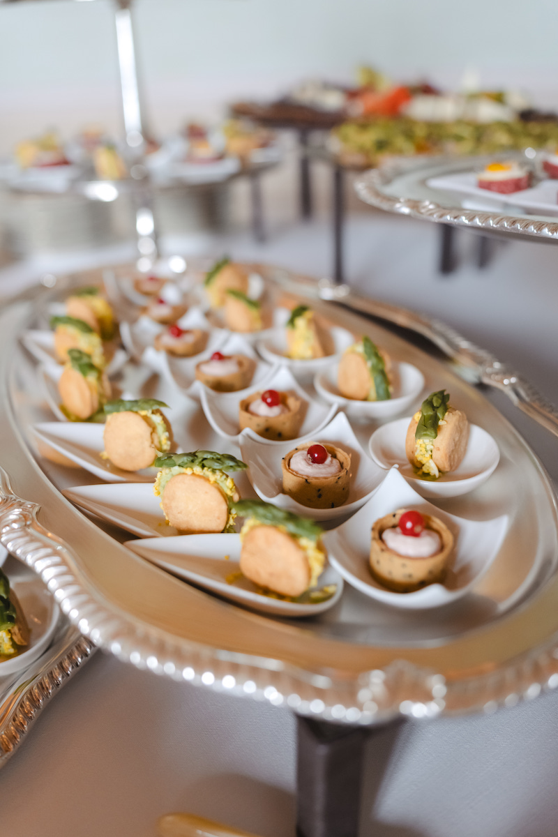 A silver tray displays assorted bite-sized appetizers on individual spoons, each topped with garnishes. More trays with similar small dishes are visible in the background.