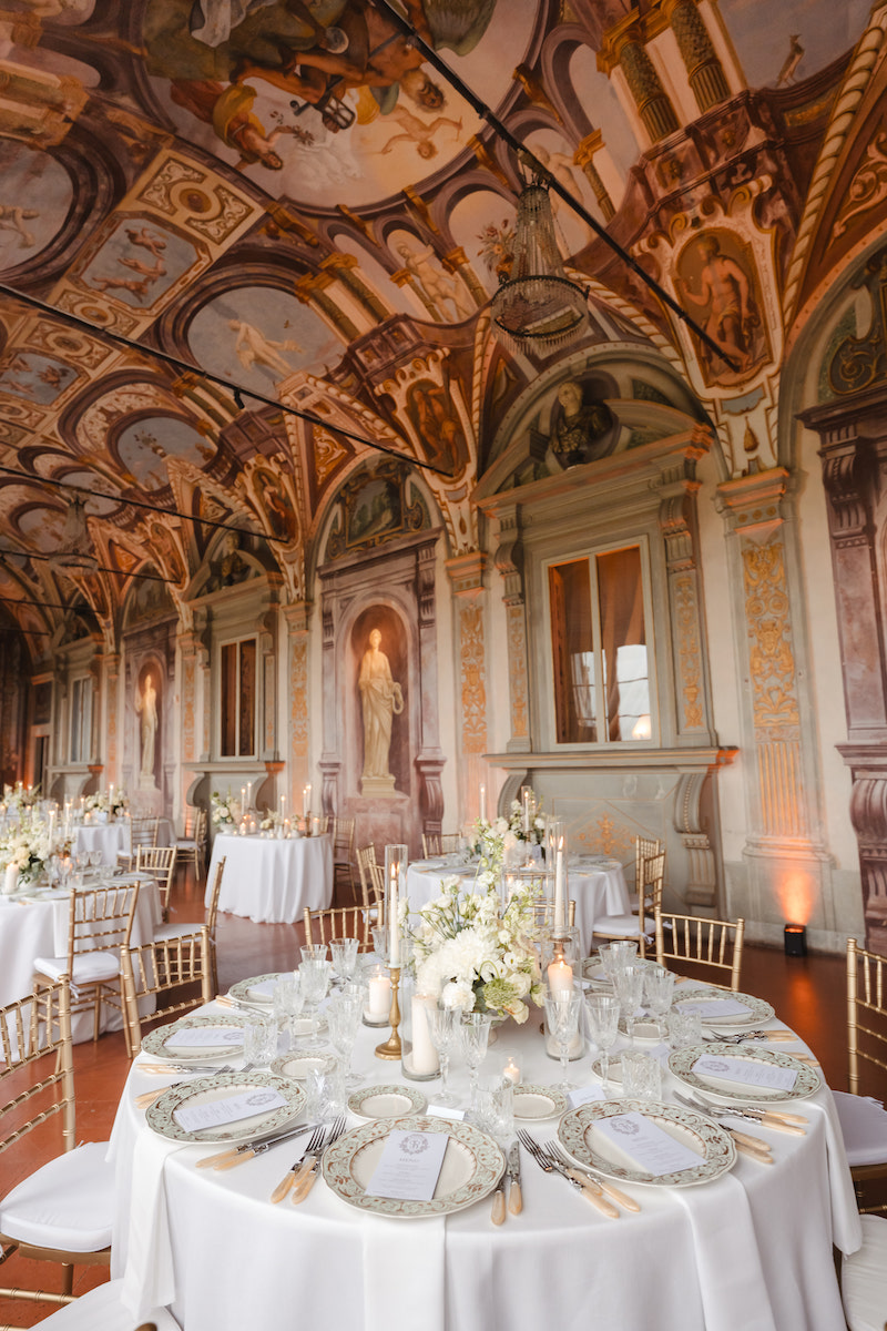 Elegant banquet hall with ornate painted ceiling, arched columns, and round tables set with white linens, floral centerpieces, and formal place settings.