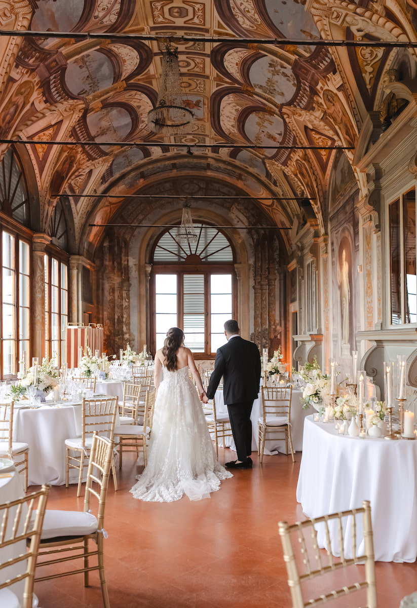 A bride and groom walk hand in hand through an ornate, high-ceilinged ballroom decorated with white flowers and gold chairs for a wedding reception.