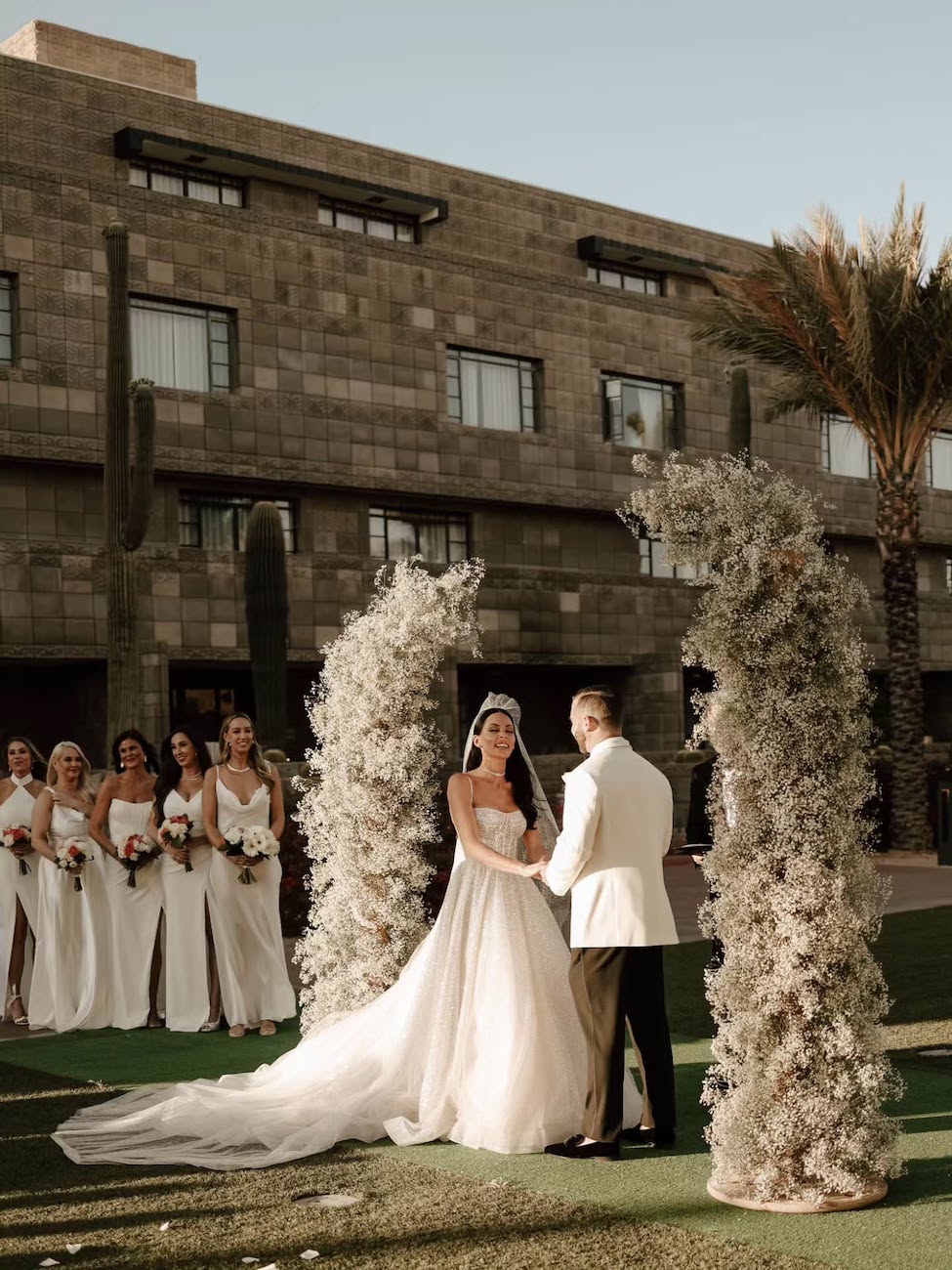 A bride and groom stand under a floral arch, holding hands during an outdoor wedding ceremony, with bridesmaids in the background.