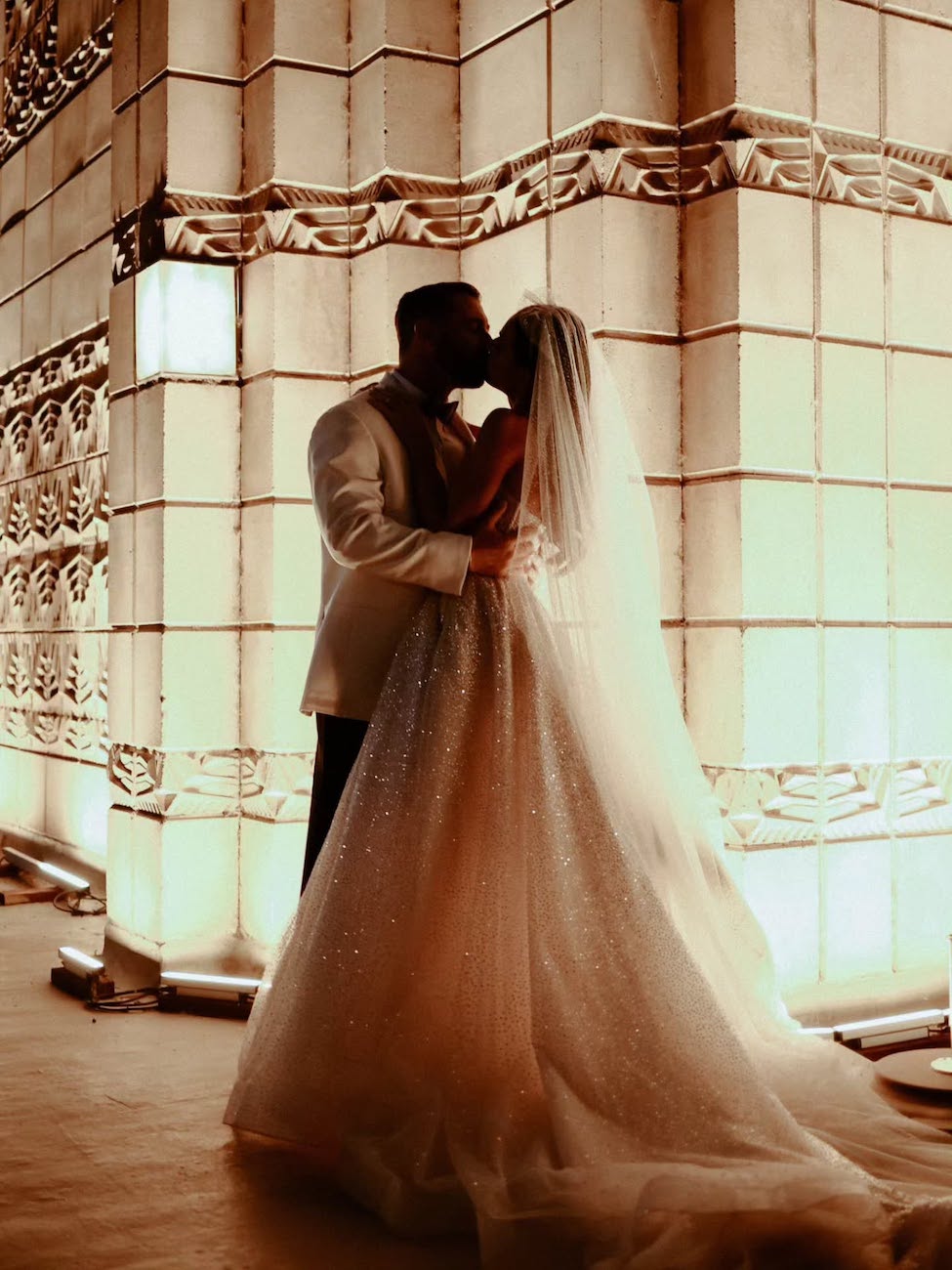 A bride and groom share a kiss while standing in front of an illuminated, ornately patterned wall, dressed in formal wedding attire.
