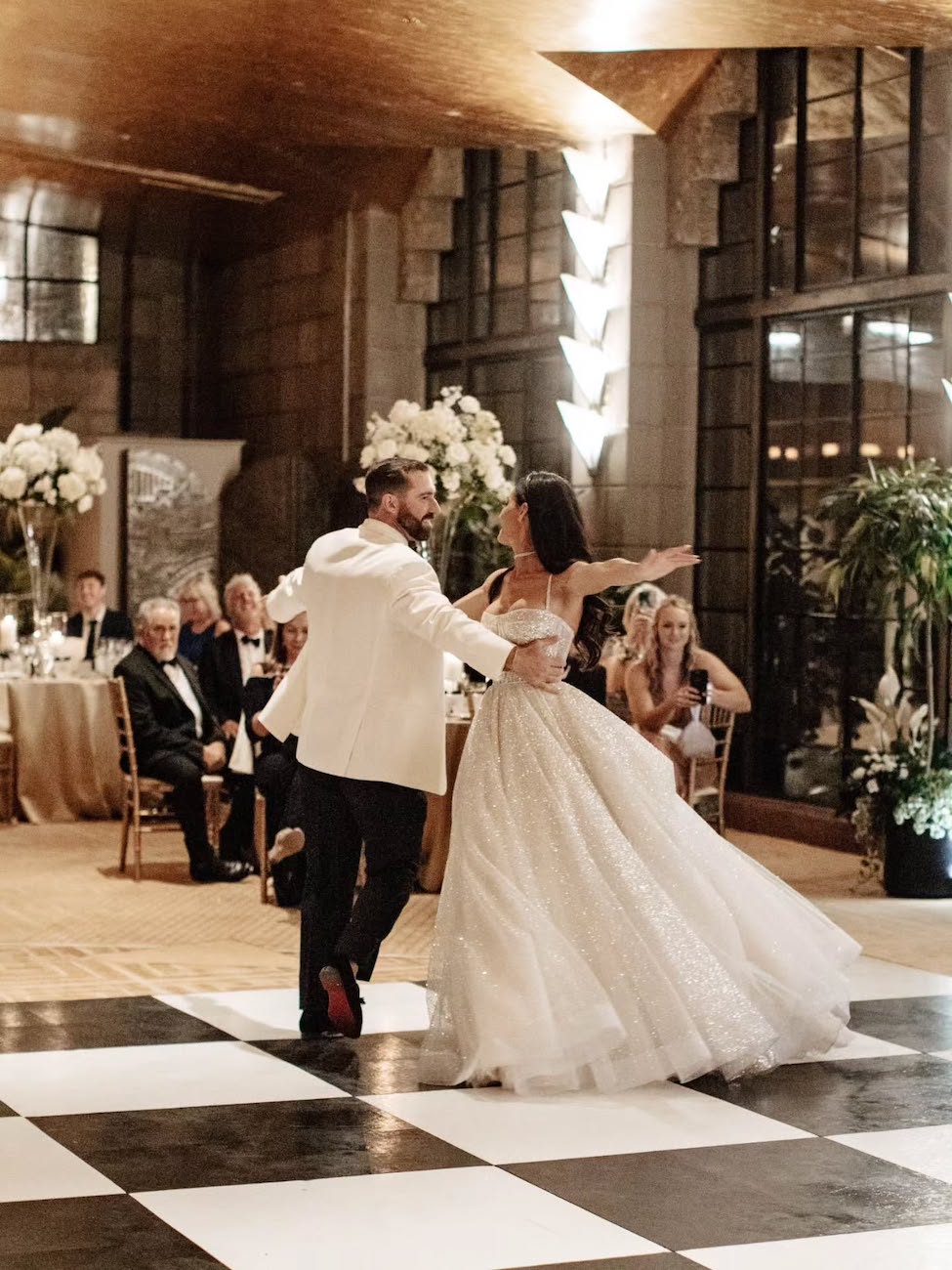 A bride and groom dance on a black and white checkered floor at their wedding reception, with guests seated at round tables in the background.