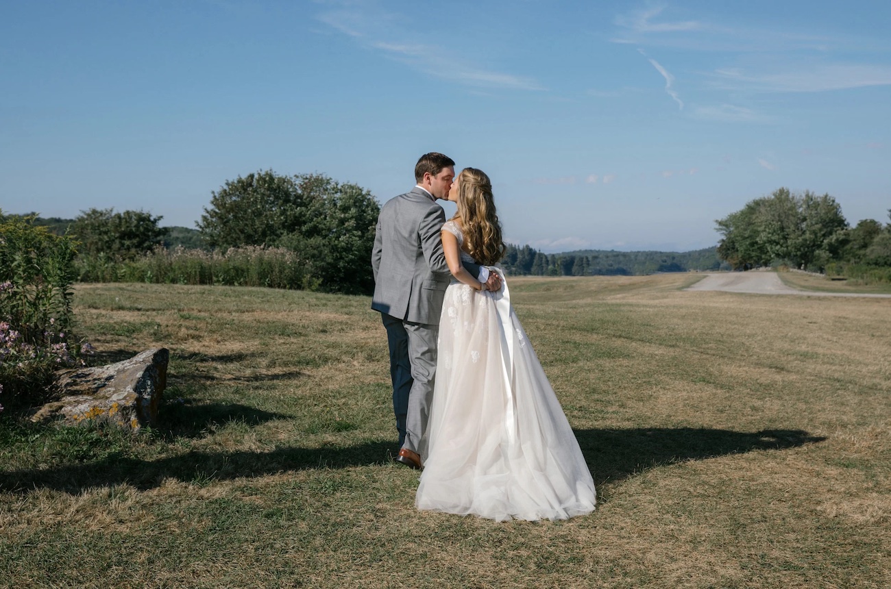 A bride in a white dress and a groom in a gray suit walk together on a grassy field under a clear blue sky.