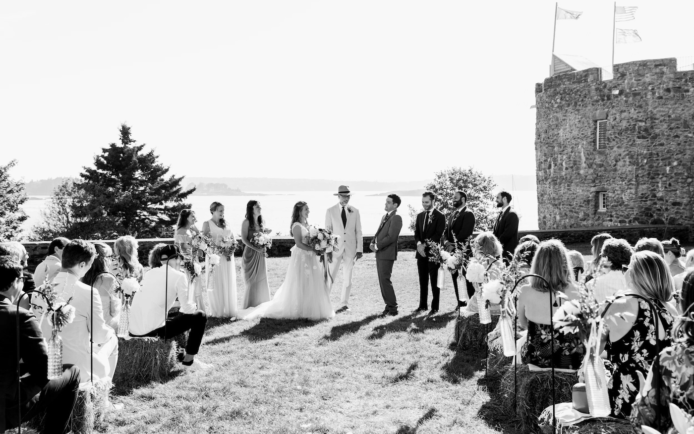 A wedding ceremony takes place outdoors, with the bride and groom facing each other, attendants standing by, and guests seated on either side.