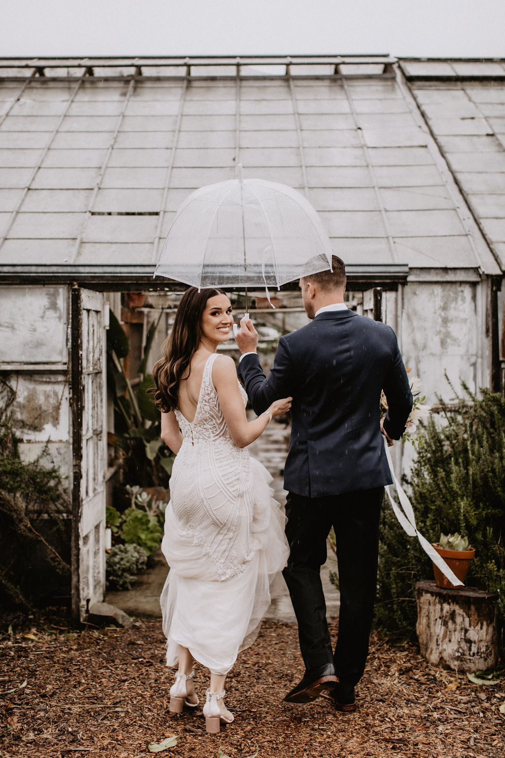 A bride and groom walk outside under a clear umbrella, with the bride looking back and smiling near an old greenhouse on a cloudy day.