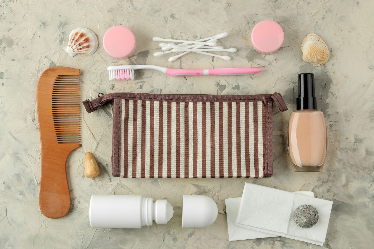 A striped toiletry bag surrounded by a comb, cotton swabs, toothbrush, nail polish, deodorant, lotion, cotton pads, and seashells on a gray surface.