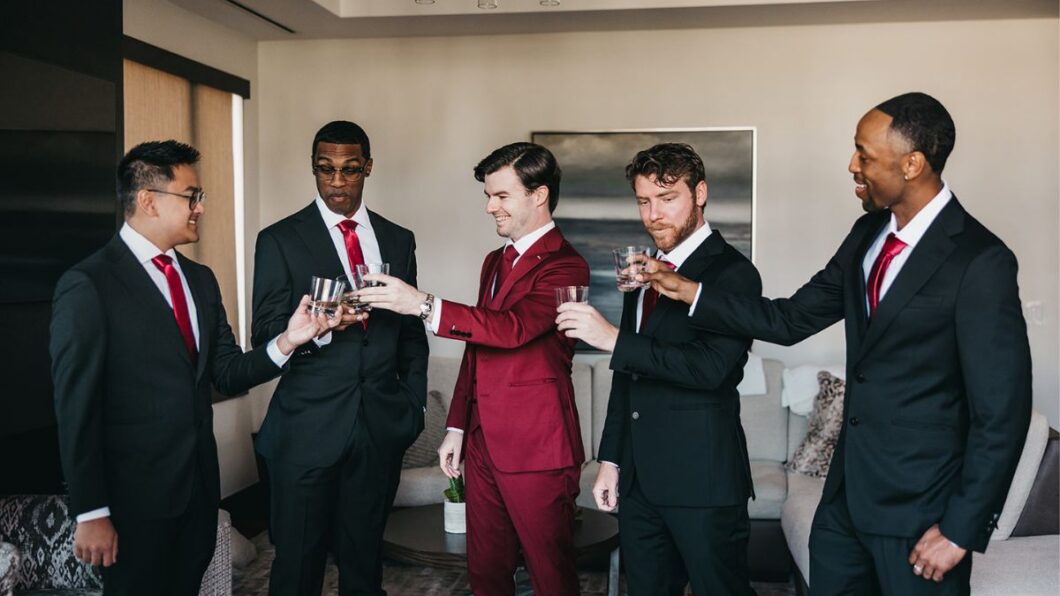 Five men in suits, one in a red suit and four in black suits, stand in a living room toasting with glasses.