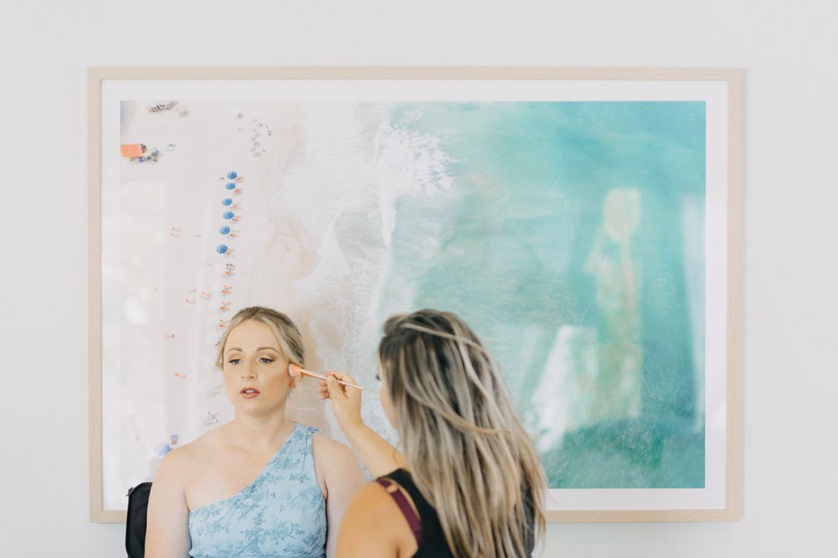 A woman applies makeup to another woman sitting in front of a large, abstract painting with blue and green tones.