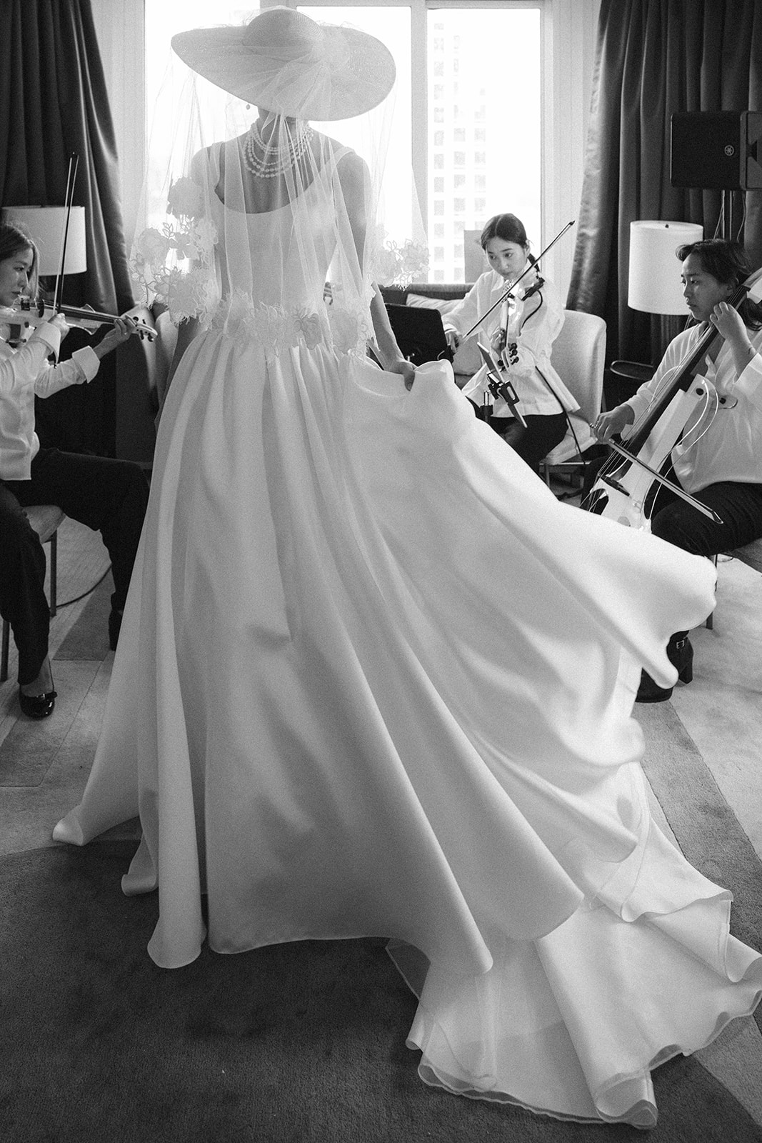 A woman in a bridal gown and wide-brimmed hat standing in front of a seated string quartet.