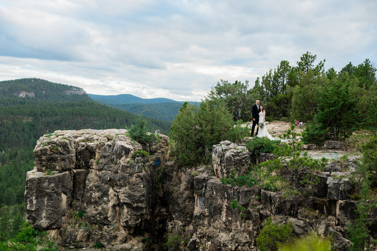 A bride and groom stand on the edge of a rocky cliff overlooking a forested valley.