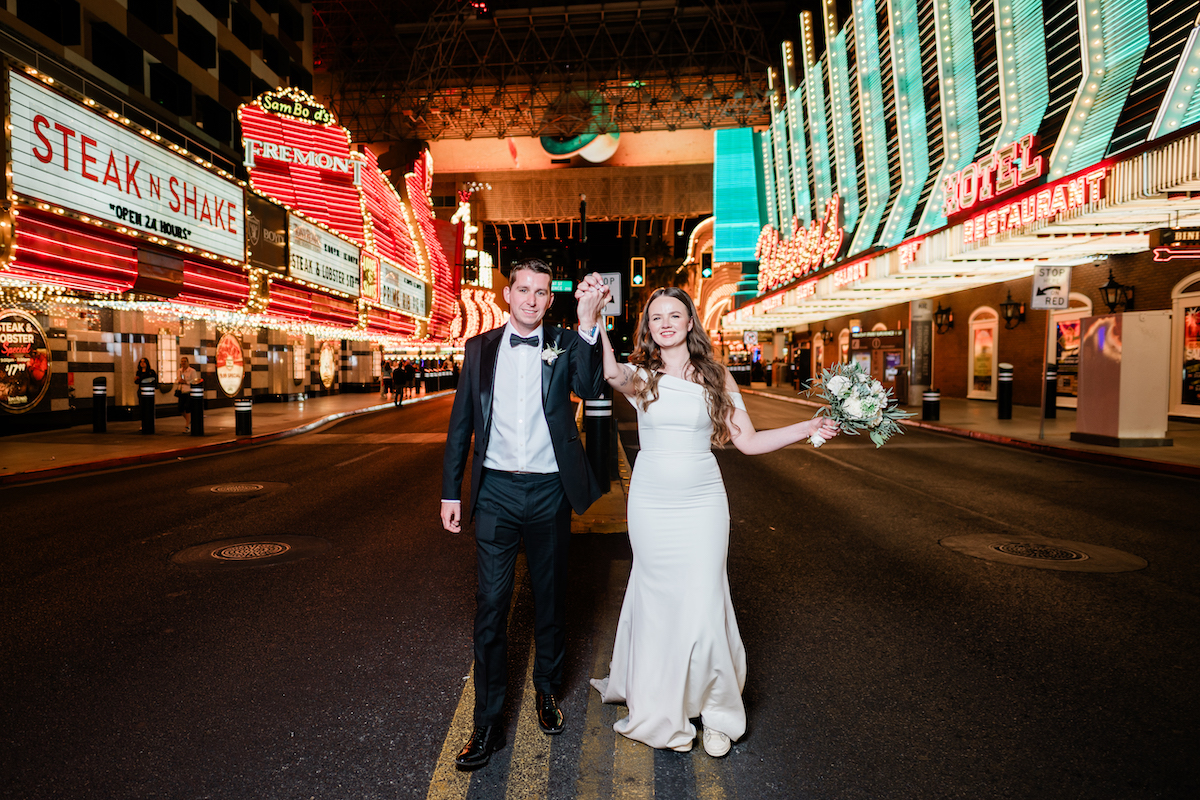 A bride and groom holding hands and walking down a deserted street in Las Vegas lined with illuminated neon casino signs.