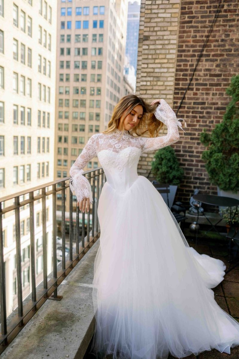 A woman stands on a balcony wearing a long-sleeved white lace wedding dress with a full tulle skirt.