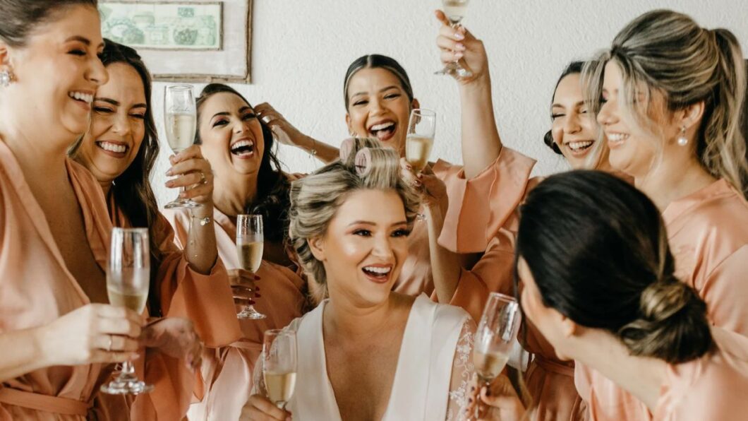 A bride with hair rollers in her hair sits surrounded by smiling bridesmaids holding glasses of champagne.