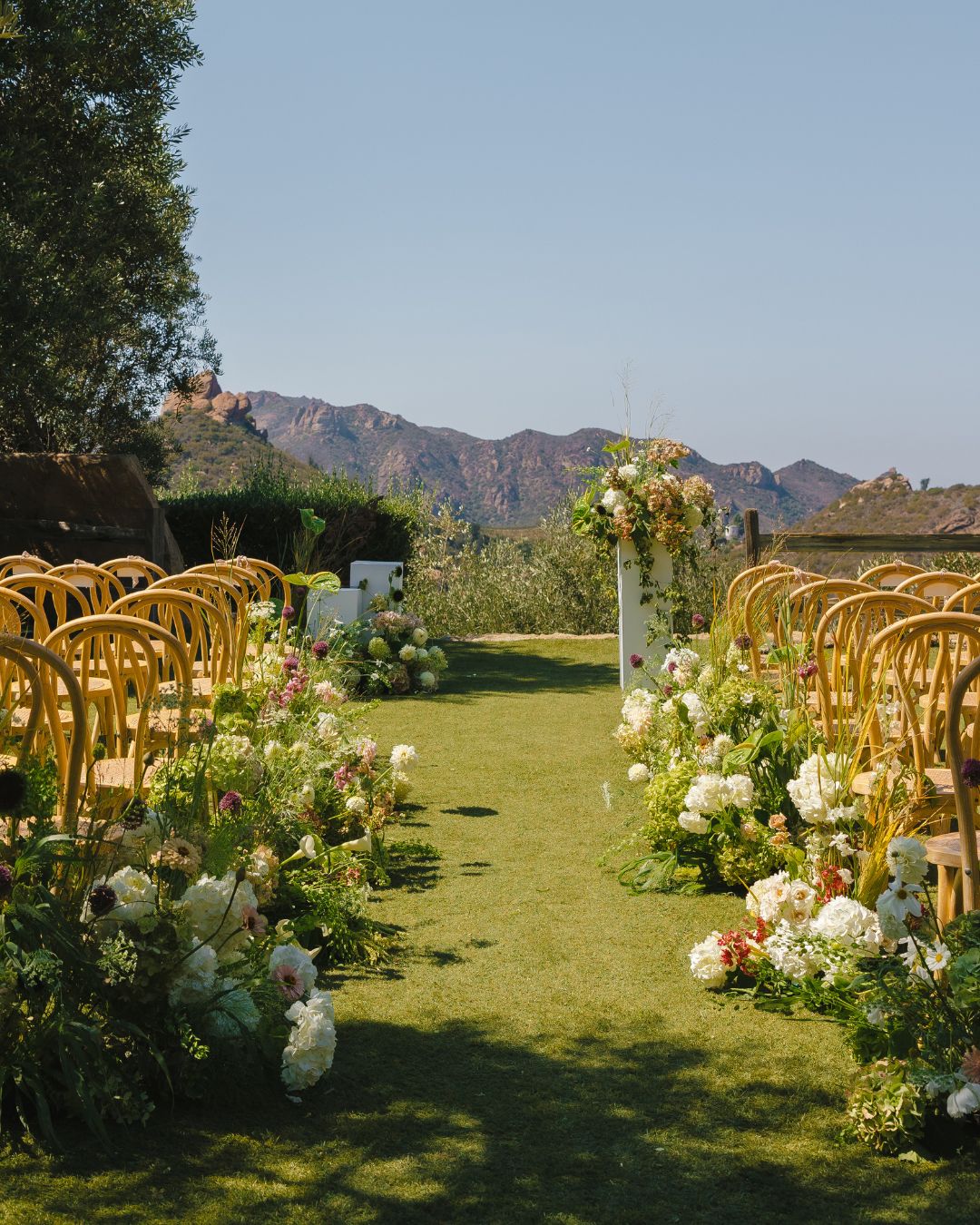 Outdoor wedding ceremony setup with rows of wooden chairs lining an aisle decorated with floral arrangements, overlooking a mountain landscape.
