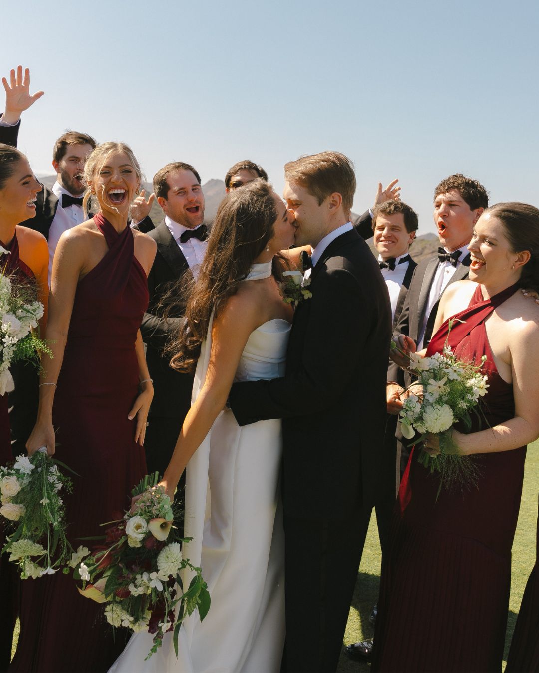 A bride and groom kissing, surrounded by smiling bridesmaids in burgundy dresses and groomsmen in black tuxedos.
