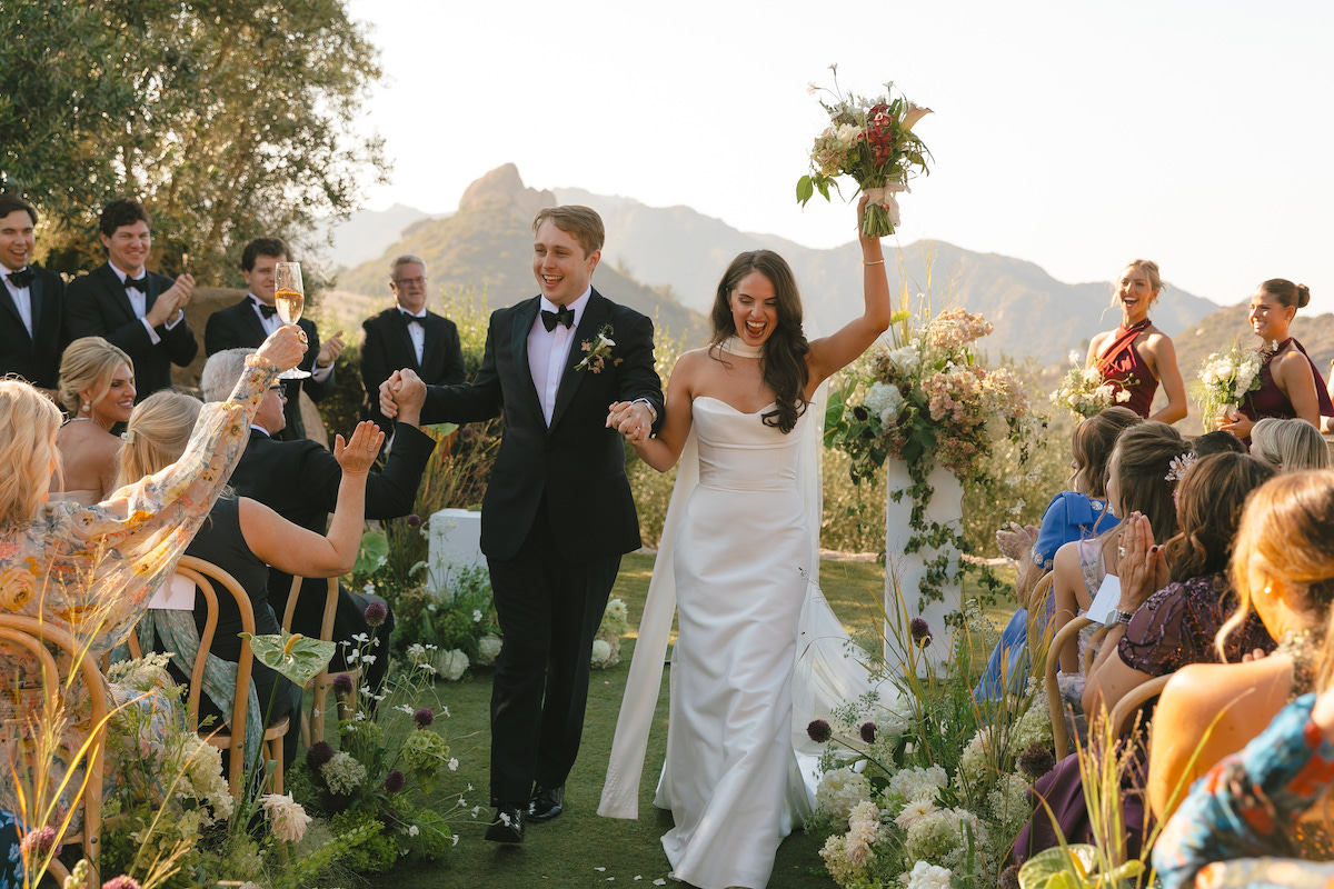 A bride and groom walk hand-in-hand down an outdoor wedding aisle, holding hands and smiling toward guests.