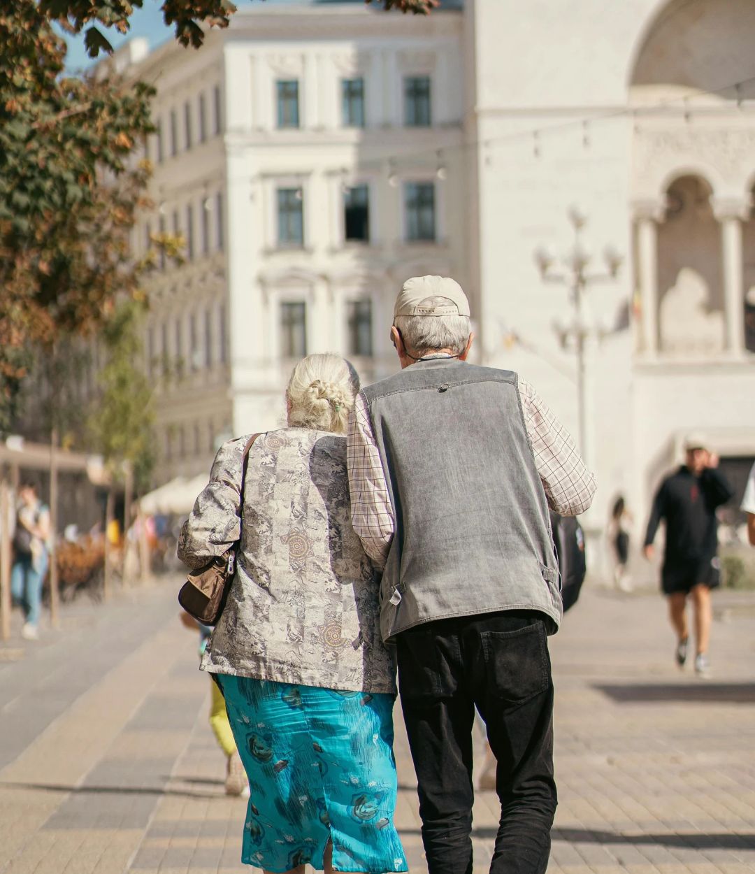 An older couple walking arm in arm through a sunny city square.