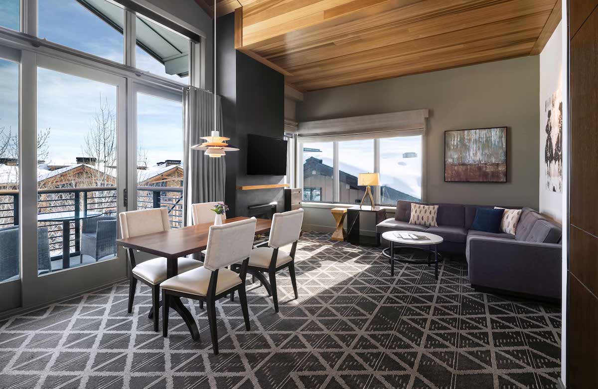 A modern hotel suite featuring a dining table with four chairs, a sectional sofa, a television, and large windows with views of snowy outdoors.