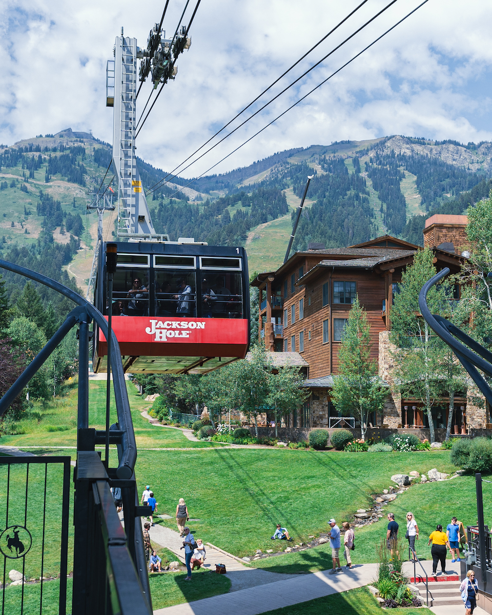 A red Jackson Hole aerial tram car moving along cables above a grassy resort area and mountain lodge.