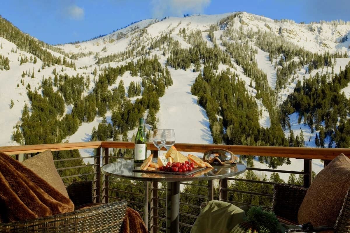 A glass table with a bottle of wine and a platter of food on a balcony overlooking a snow-covered mountain.