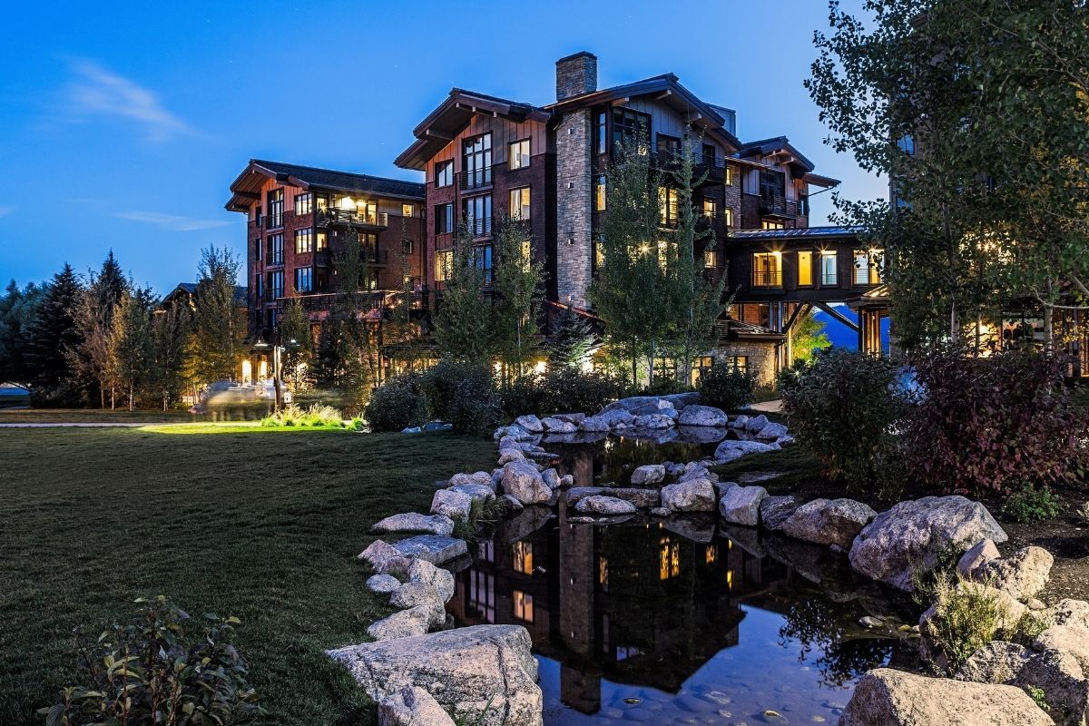 A multi-story timber lodge at dusk, with a landscaped lawn and rock-lined pond in the foreground.